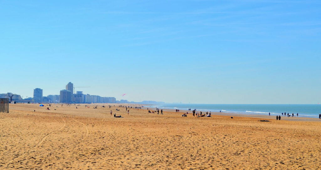 the view of the belgian coast in oostend. People are far off in the distance, little specs along the sand. Even farther are some buildings, but none too tall and they're kind of hazy as they blend into the bright blue, completely clear sky.