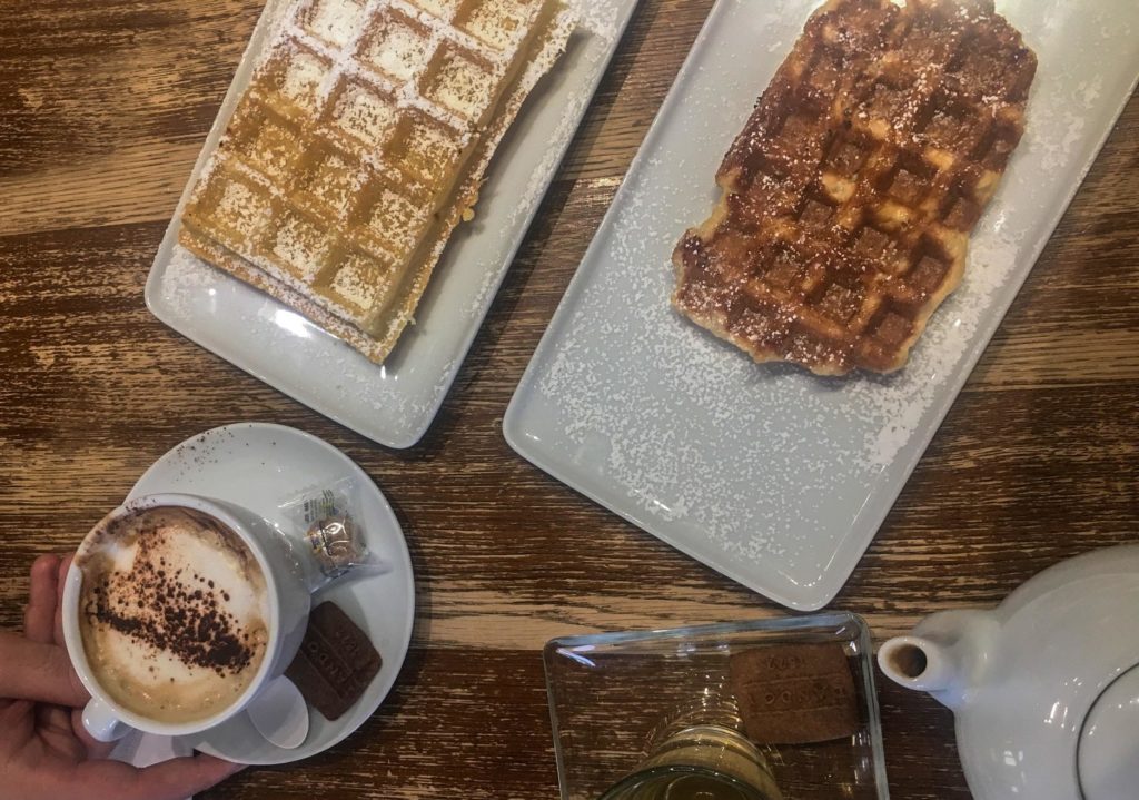 two waffles from above with a cappuccino and a pot of tea from Maison Dandoy. 

One rectangle waffle is lightly golden with powdered sugar on top.

The second waffle is much darker golden brown, and not as perfect, it has uneven edges - this is the liege waffle. It has less powdered sugar on top.