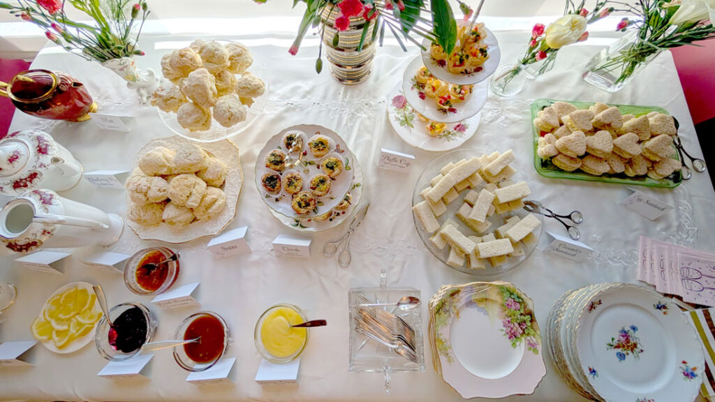 an overhead photo of a beautifully set afternoon tea party table. There's a white on white sewn  tablecloth with pink flowers in vases, three tea pots, two plates of scones, two tiered plates with savoury pastries, two plates filled with tea party sandwiches, a stack of empty plates, four small bowls with jams and sauces for the scones, a plate of lemons. 