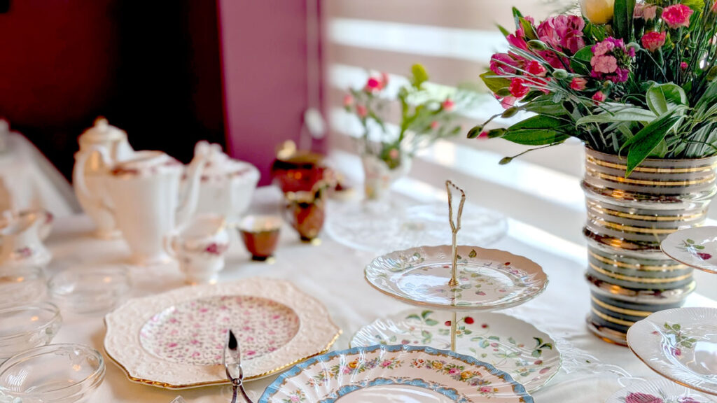 the view of the set up of an afternoon tea table with empty tiered plate, empty serving trays, and teapots. There are also a large vase filled with a bouquet of flowers on the table. 