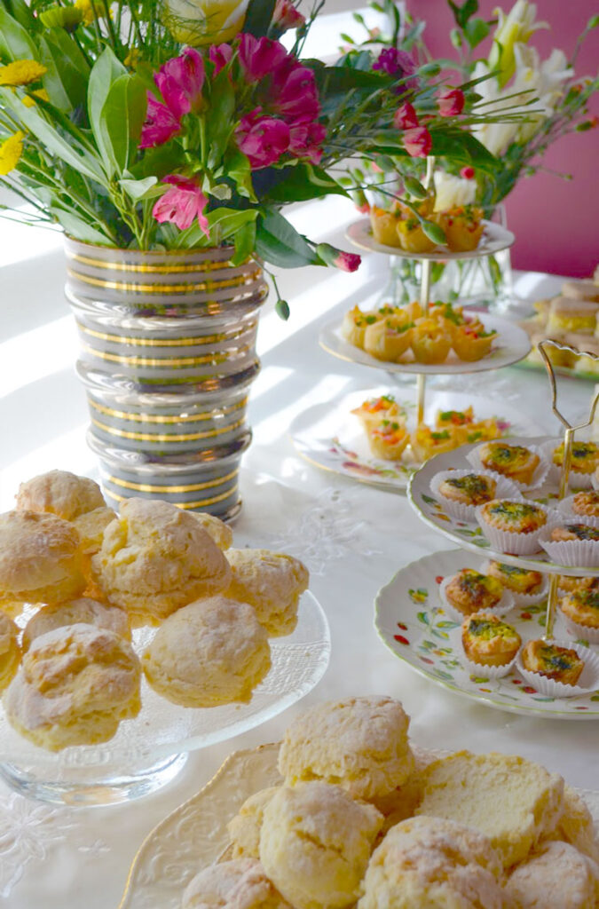 a table set up for afternoon tea, with a white tablecloth underneath, two plates (one raised) filled with large scones, a two and three tiered plate with mini quiches and savoury pastries and to bring it all together a vase with a large bouquet of colourful and lively flowers. 