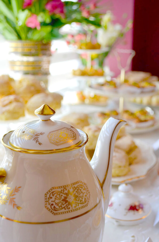 a close up of a beautiful tea pot that is glazed shiny white with gold decals on it. Behind it is a blurry, but well set up afternoon tea party table. There are scones, tiered plates, and flowers.