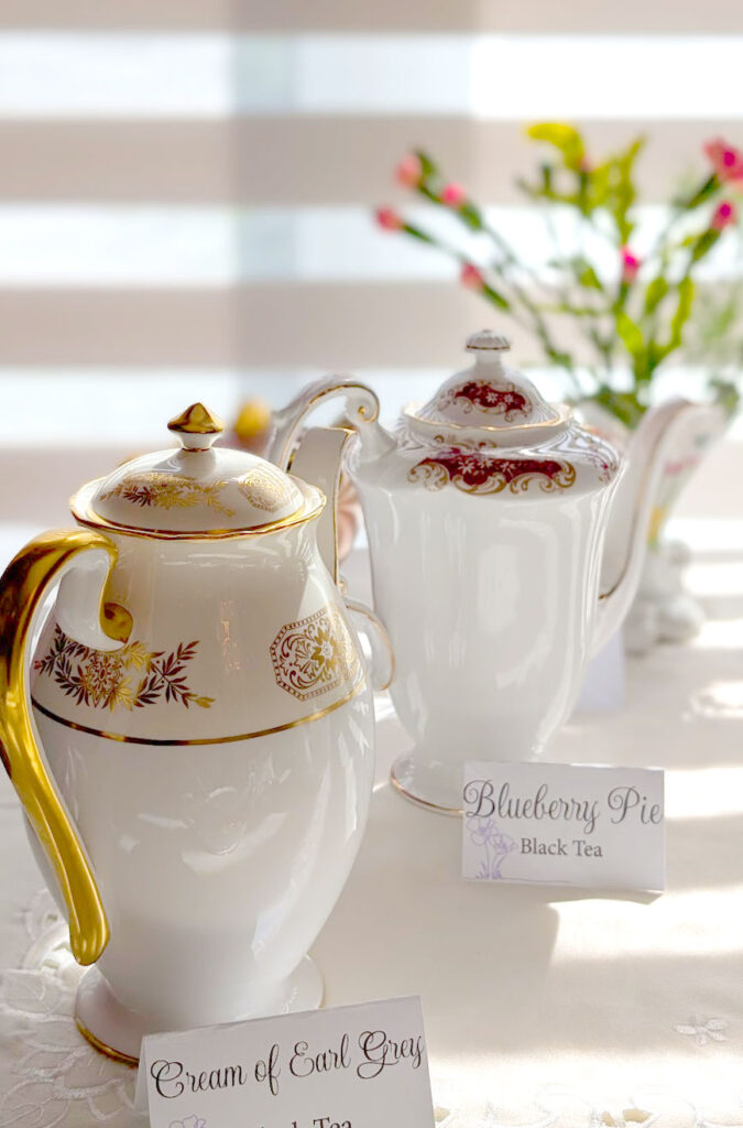 two tall teapots serving tea at afternoon tea. The one in front is white with a gold pattern, there's a sing in front that says Cream of Earl Grey, Black tea. Behind it is a white with a burgundy pattern on the top teapot. This one has a sign that says Blueberry Pie, Black Tea