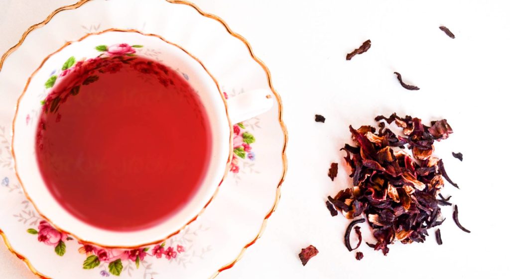 benefits and risks from hibiscus tea featured showing an overhead photo of a delicate white with pink and red flower accents and a painted gold rim around the plate underneath a teacup. Filled almost to the brim is a deep red tea liquor. Next to the saucer, on a white background are dried red hibiscus petals in a pile.