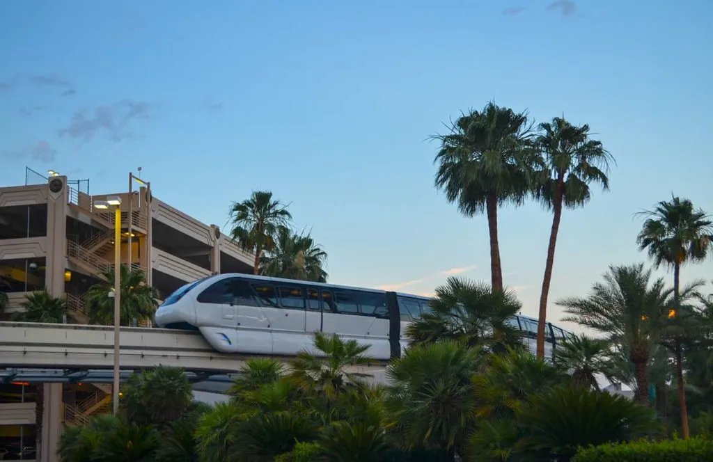 take the free monrail to when visiting vegas on a budget. There's an elevated railway with a white tram speeding across it. It loos futuristic with white and completely dark black windows so you can't see inside. There are palm trees all around it and a parking garage behind it. 