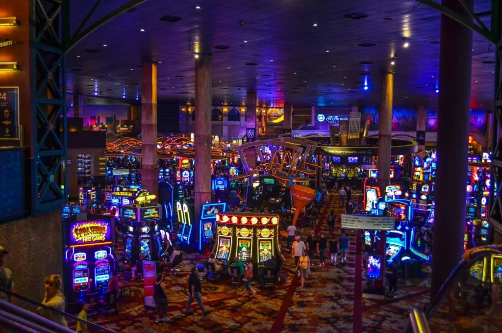 money saving vegas casino room. It's a dark room with a patterned carpets and zero windows. You see lines of neon lit slot machines at the front of the room but by the back of the room they're all blended together in a wave of neon lights. 