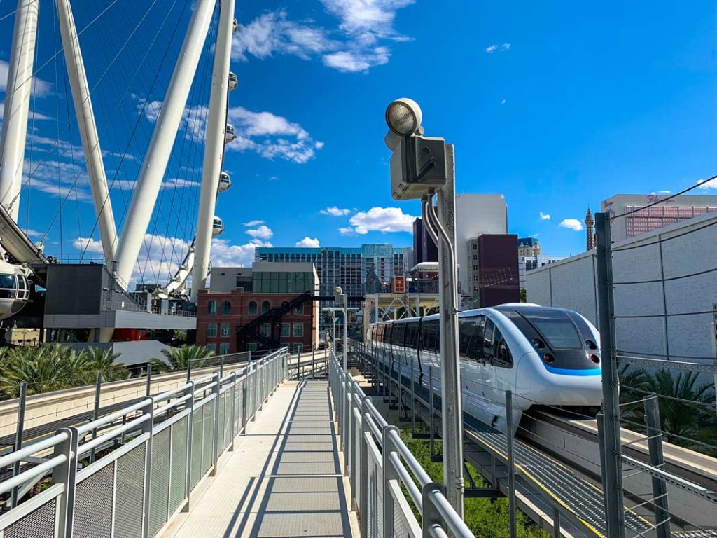 save money in las vegas on the free monorail. There are two tracks, on the right track a monorail just left. It's going towards the buildings of vegas, and the giant ferris wheel going high into the bright blue sky
