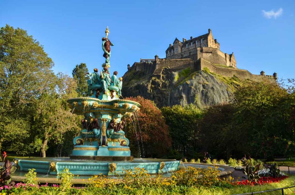 princes street gardens in Edinburgh Scotland. It's a bright sunny day, with the tiniest cloud hovering in the sky on the top right. Towering above everything is, a giant rock formation  and a castle built on top of it with a stone wall all around for extra protection. Trees line the bottom of the mountainous rock, bringing your eye to the front of the image where a baby blue and gold found sits. It's actually the same height as the castle in the background, but that's just the perspective. Flowers and bushes circle the fountain at the bottom. 