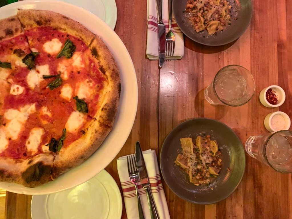 wolfgang puck sharing food vegas hack. An photograph from above showing a full margarita pizza with fresh basil leaves, and two small appetizers on a wooden table with cutlery on top of fabric napkins