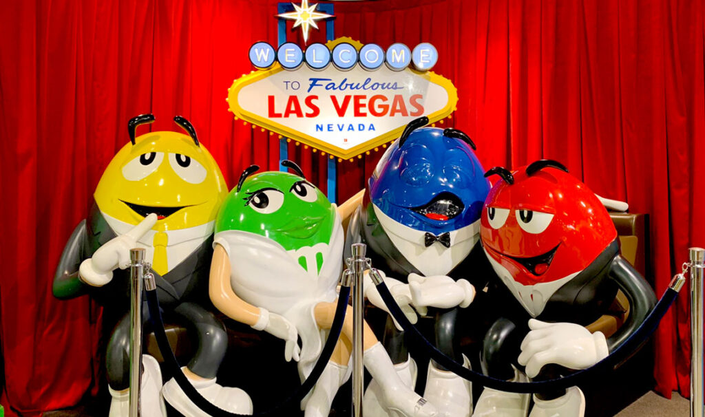 a red curtain in the background with the iconic welcome to las vegas sign hanging from the ceiling behind the four life size statues of M&M personas: the yellow, green, blue, and red MnMs