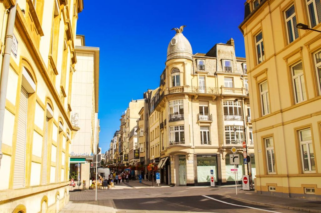 an empty luxembourg city street. It's looks like a yellow and blue picture only. The sky is only blue and the stone buildings are a yellow stone.  You can see people are walking further up ahead in the street