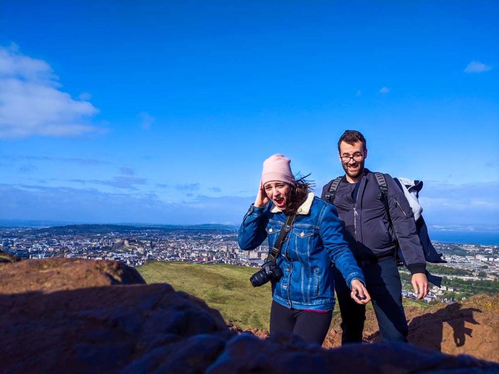 a couple stands a top the insanely windy Arthurs seat in Scotland. They are on a rocky surface and the city of Edinburgh is very small buildings lining the background. You can see some slight hills, and water behind the city before the sky takes over. 

You can tell it's window because the woman is holding on to her pink beanie hat, her hair is blowing in the wind. Her mouth is wide open in shock. She's wearing a completely buttoned up sherpa lined jean jacket. There's a camera hanging cross body around her waist. 

Just slightly behind her stands a man with an unhappy face. His bomber jacket is puffed up from the wind. And he has a wide stance to stay steady so that the window doesn't push him. 