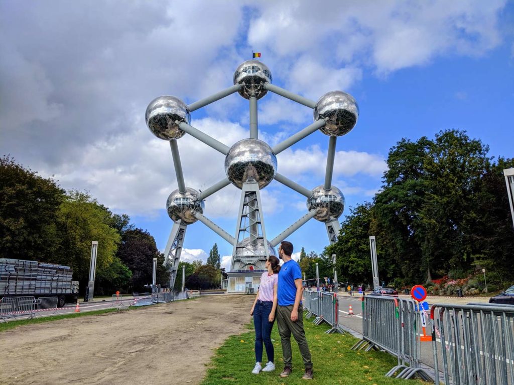 a couple stand in t shirts and pants looking behind them at the grand atomium of belgium. It looks like a silver atom, with six connected balls hovering in the air.