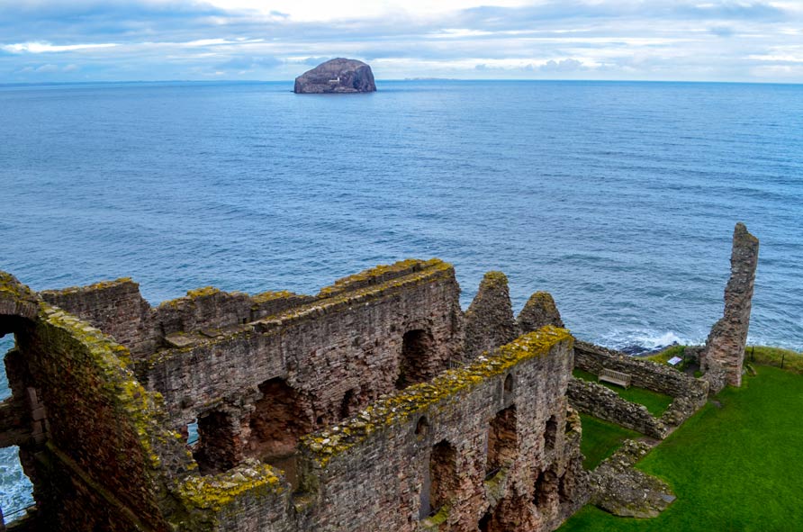 The view from the top tower of tantallon castle in Scotland, north berwick. You can see castle rows, about three incomplete stories of ruined walls below you. The stones and bricks are uneven, a mix between grey and red tints. On top of the walls, you can see moss growing. The ruins are on the edge of land, behind it the sea begins, it looks choppy as it hits the cliff wall. The  only thing in the water, in the distance, almost at the horizon is a lonely rock. You know this is bass rock, you can barely make out a small white building in the middle of the rock.