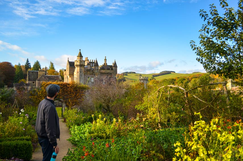 a man in scottish tweed hat stands on the side, looking back at abbotsford house in the scottish borders. IT's tucket behind lush gardens, and before the rolling mountains. It looks more like a castle than a house, with spikes and towers built in a beautiful stone. 