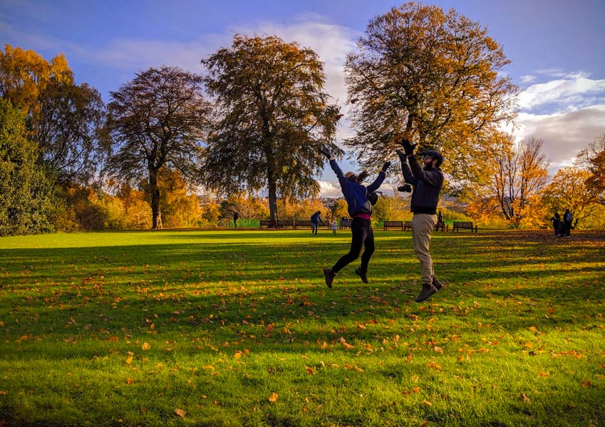 two people are jumping for joy in a field in the royal botanical gardens in Edinburgh in the fall. The trees behind them are starting to change colours from green to burnt yellow. There are some leaves below them on the grass that have already fallen. The couple is also dressed for fall, wearing a fall jacket, boots, a hat, and gloves. 