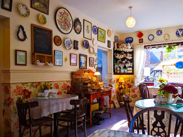 inside Clarindas tea room, there are plates and picture frames on the wall with wall paper on the bottom half.It has old style furniture that you would see in your grandparents place