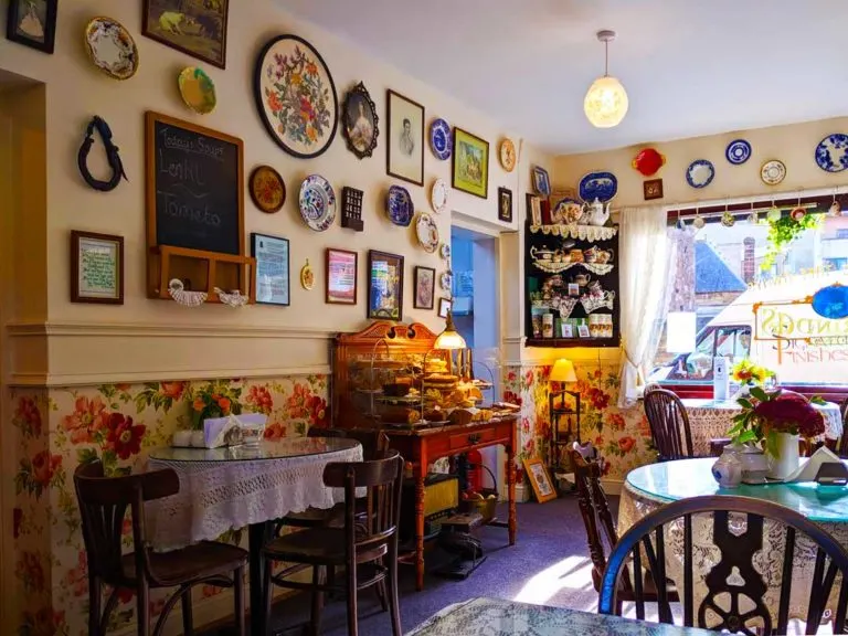 inside Clarindas tea room, there are plates and picture frames on the wall with wall paper on the bottom half.It has old style furniture that you would see in your grandparents place