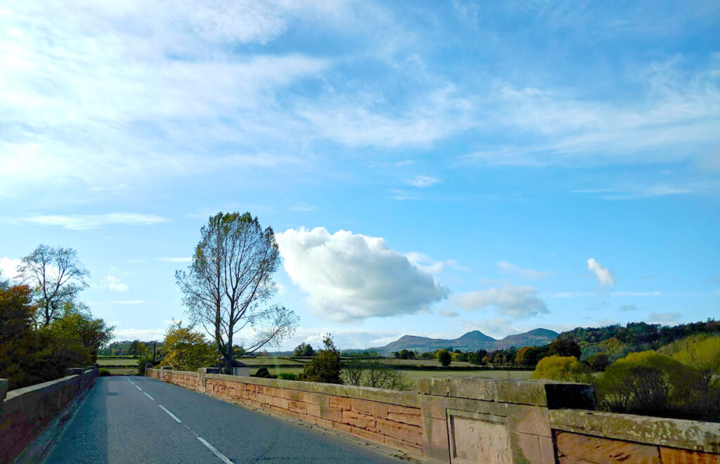 driving through the scottish borders. A two lane street on a bridge, both sides are lined with red brick stones, there isn't a lot of room for error. Beyond the bridge is green farmland, a tall tree without any leaves, and far off in the distance are some mountains. 