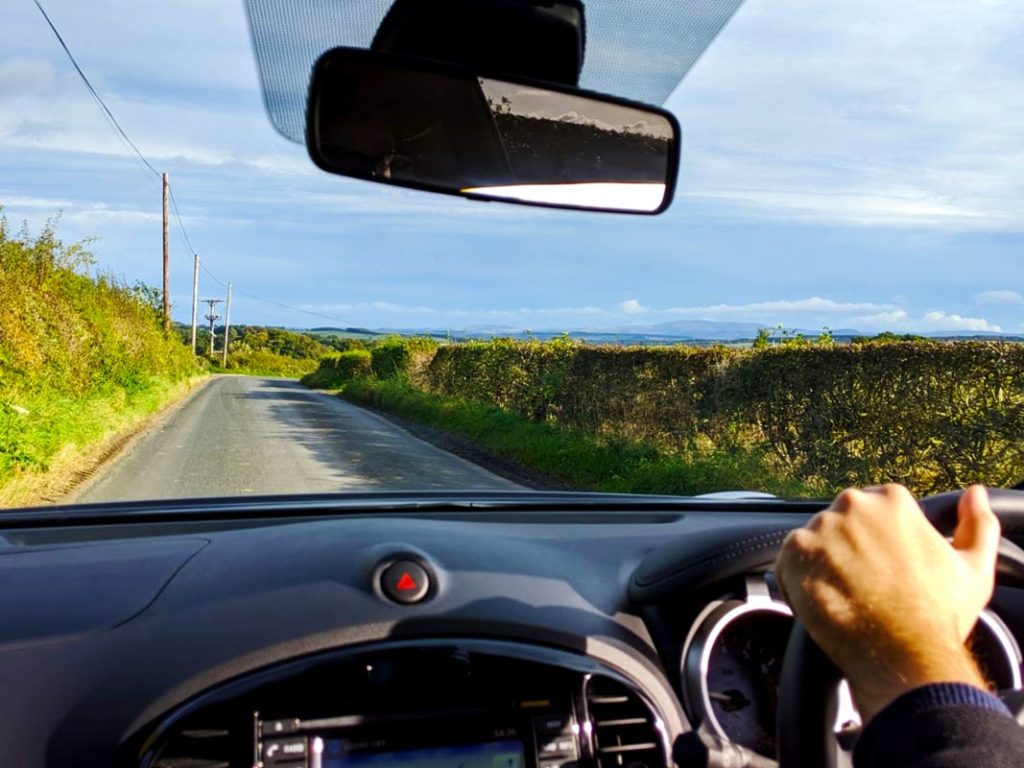 the view of the road through the windshield of a car. You can see the dashboard, and a mans hand grips the wheel (on the right side of the car). The road is narrow, only fitting one car as there's tall grass on either side.