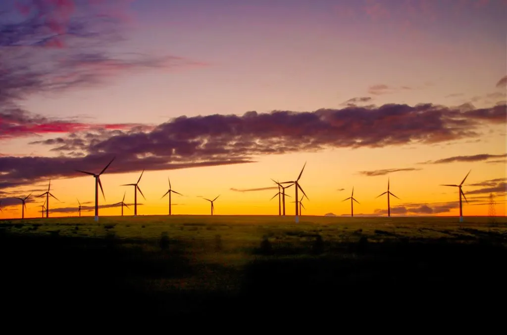 a magical sunset in scotland across the farmland populated with solar wind fans. The sky goes from a deep orange on the horizon, to a rich golden yellow until turning to purple. 

The rest of the grass and farmland is in shadows, but you can almost make out a greenish tint. 