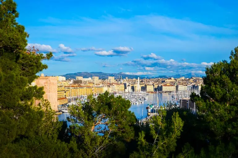 framed-marseille- through some lush green trees. You can see the port and then the city and mountains in the background with blue skies with a few clouds - is marseille dangerous