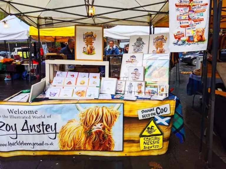 hairy-coo-artwork roy anstey booth at the market in edinburgh banner on the bottom of a table with his printed artwork on the set up on display