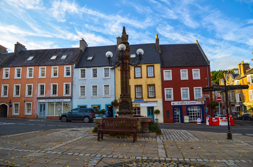 jedburgh town in the scottish borders. You stand in the middle square of the town, with a stone monument and light fixture in the middle. Behind the monument is the road and four attached homes lining the street, each one is three stories, with a shop on the main floor, and all of them a different colour. The largest house on the left is orange, attached to blue, yellow, and red homes.