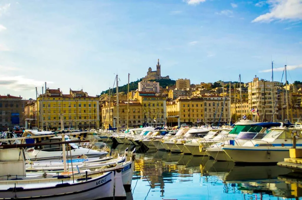 what marseille is like - marseille-old-port- with boats lined up all along the water on a sunny day but you can still see their reflection in the very still water. 

Yellow apartment buildings are lining the background. and Towering above everything is the basilica on a lush green mountain