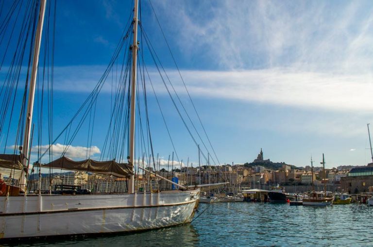 marseille-port-and-basilca-views: a large sailboat in the front on the water and you see the shadow of the basilica in the background hight on the mountain