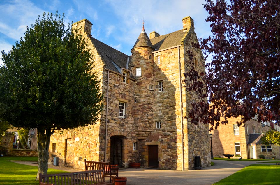 the outsode of mary queen of scotts exhibit. It's an old stone building, with different sizes and colours. It's about 5 stories with randomly offset small window. There's a narrow tower in the middle of the house. There's also a tree on either side of the house.