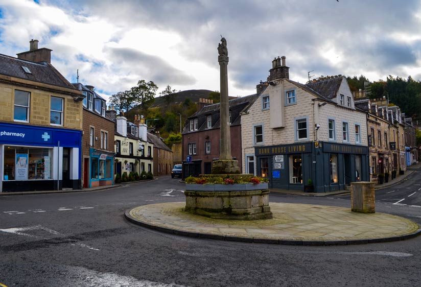 the view of melrose in Scotland. This is a roundabout in the middle of the town, connection at least two streets. There are homes lining the streets, very small town vibe homes with only three stories, but a shop is on the main floor. 