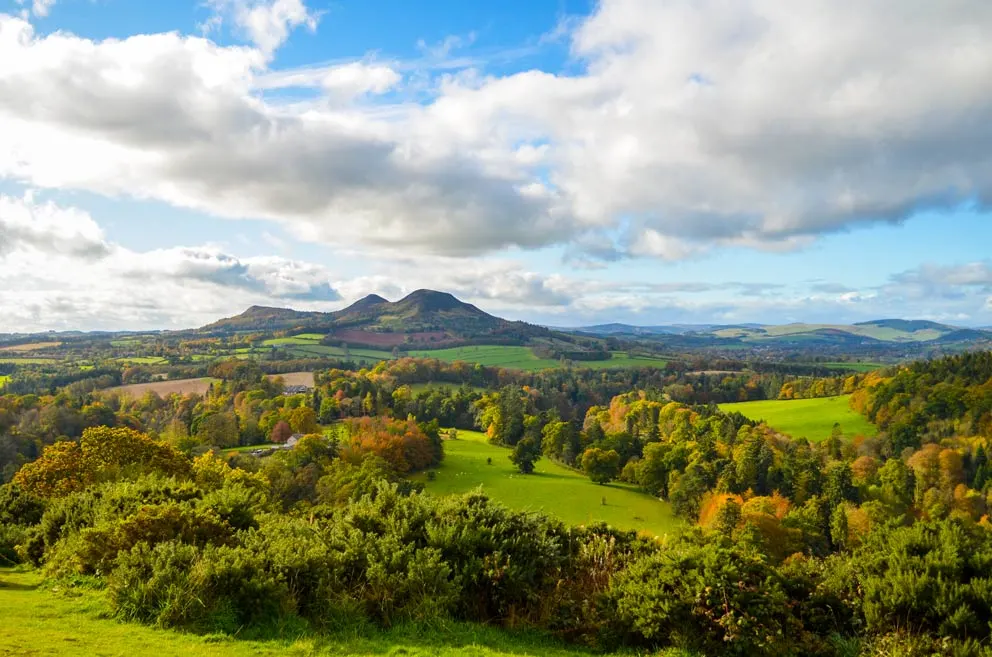 scotts view in the scottish borders. It's a gorgeous landcsape view on a cloudy, but still sunny day. The clouds look as though they have movement, large ones cover the blue sky in the foreground, but become thinner and smaller the further they are in the distance. Meeting the clouds in the distance, is a mountain with three peaks . In front of it is farm land, some covered with trees leading all the way back to the front of the image. 