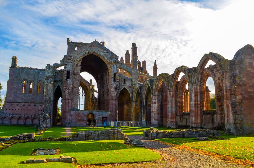 melrose abbey in the Scottish borders. Although there's no ceiling, you can see the size that the abbey once was, because the walls are still stnading. It's in the shape of a cross, with arched walls leading to the centre and an even taller arched wall in the centre. The sun is still rising, so it's peaking through all the empty archway, brightening up the green grass around the ruined abbey. 