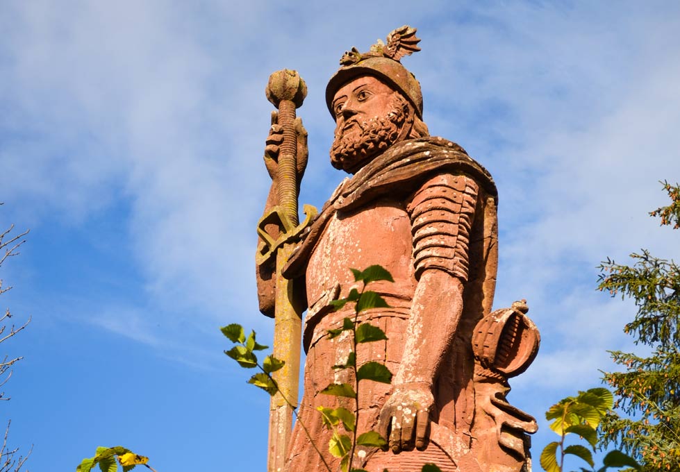 a giant red stone statue of william wallace in the scottish borders. It's a man with a beard fully dressed in armour and holding a giant staff, that's almost taller than him. You can see the edges of some trees, but the statue soars way above them, showing how tall it is!