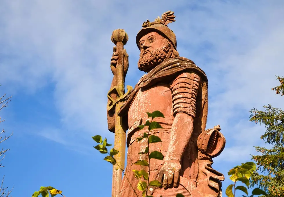 a giant red stone statue of william wallace in the scottish borders. It's a man with a beard fully dressed in armour and holding a giant staff, that's almost taller than him. You can see the edges of some trees, but the statue soars way above them, showing how tall it is!