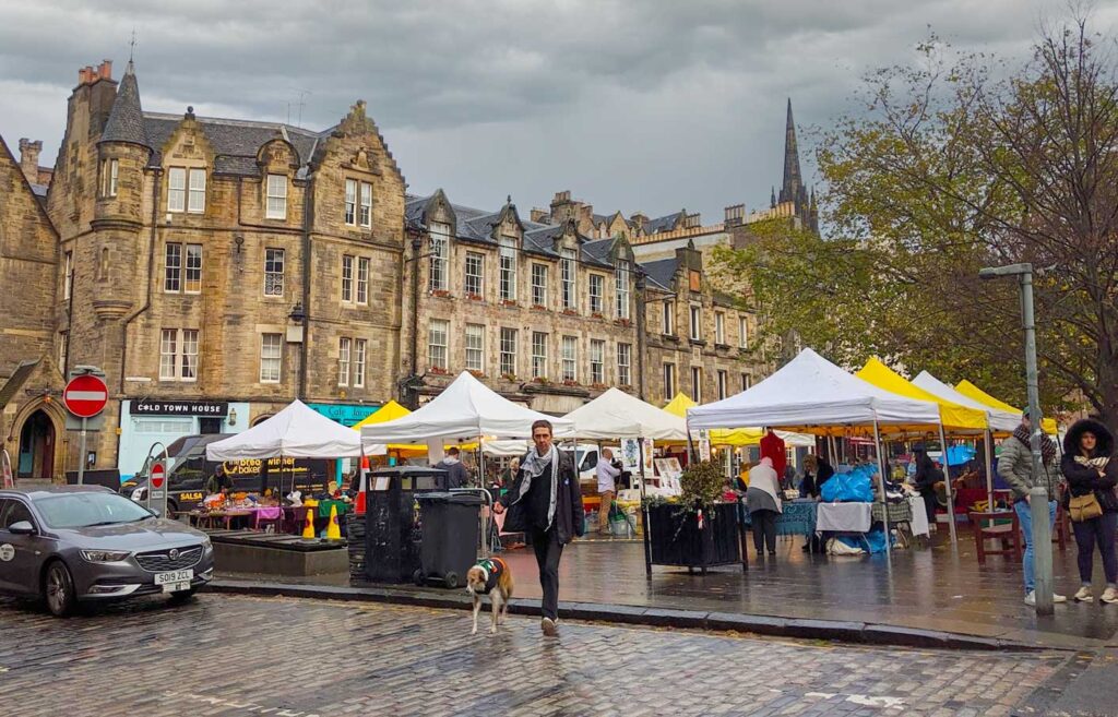 market-in-edinburgh-tents-lined-up-with-alternating-white-and-yellow-covers-old-stone-buildings-are-behind-them-people-are-walking-around-even-though-its-a-dark-and-gloomy-rainy-day