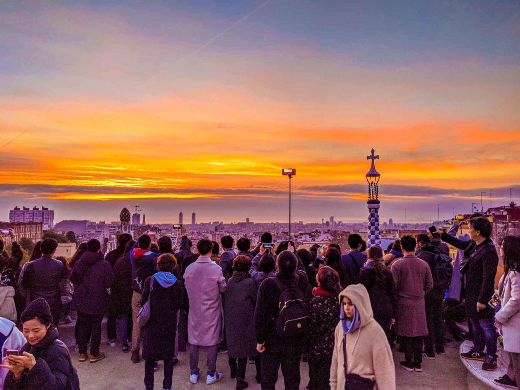 sunrise at park guell. Although the sunrise is beautiful with blue, strokes of yellow and orange and purple. But you can't see the main view of park guell, only the tower and cross as there's a crowd of people blocking the view. It's colder, a woman in front is bundled in a sweater with her hood covering her head. 