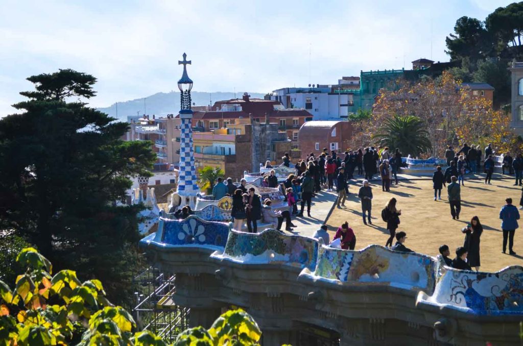 looking from outside into the restricted zone in park guell. Its the famous mosaic balcony, overhanging above the lefge. The barrier is beautiful mosaic work. People are walking on the platform. There's homes in the background and even a mountain further in the distance