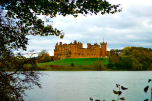 Linlithgow palace castles near Edinburgh is a reddish light brown castle sitting atop bright green grass on a slight hill. The castle is frame by leaves on the top and left side of the image. And in front of it is a lake. It's a bit of an ominous day with overcast clouds, looking like it's about to rain