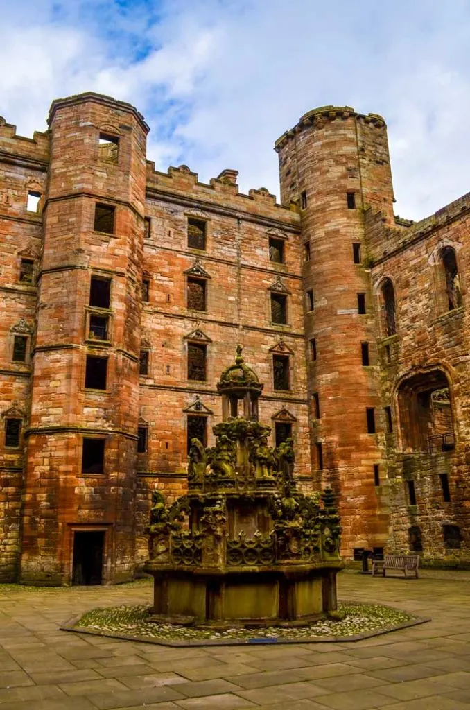 a vertical photograph of the inside a square in Linlithgow palace, showing the height of the walls, which is five stories tall. The walls area reddish brick. There are two towers that are higher than the walls (6 stories). There are a lot of empty spaces for windows. The floor is a grey stone. In the middle is a grey stone fountain, without any running water). There is some green moss growing on the fountain.  