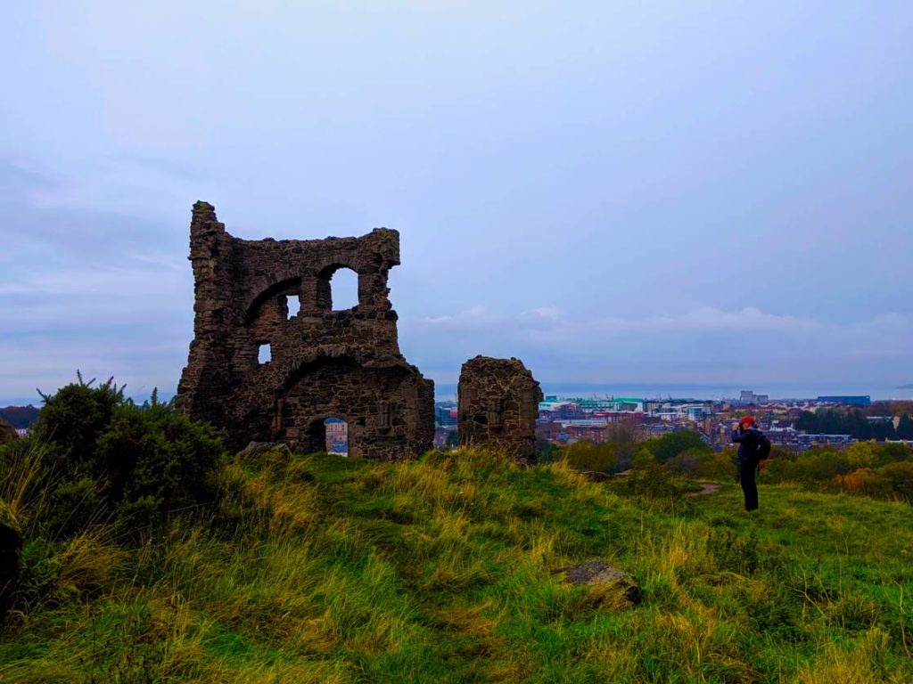 St. anthonys chapel, a random ruin, just barely still a wall, popping out from overgrown grass, with the cityscape behind it. A man is photographing the ruins. 