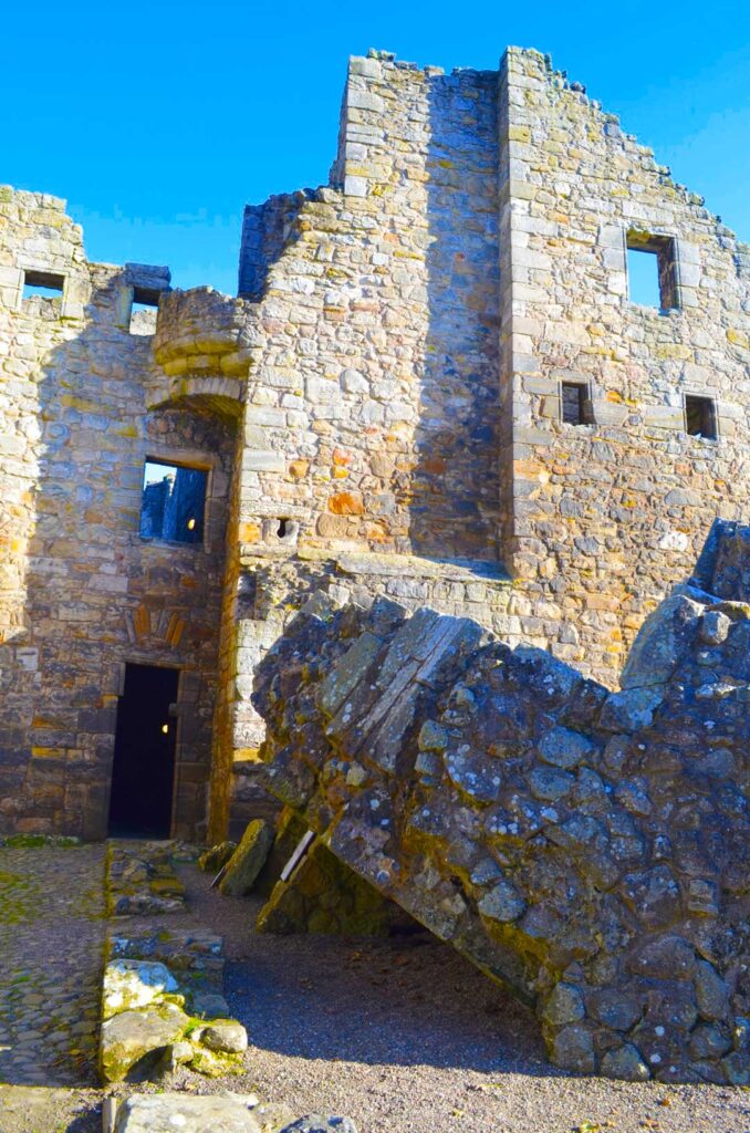 vertical image of the castle ruins at aberdour castle in scotland. A peice of the building has fallen down, there's no roof but there are three stone walls left standing with window frames. 