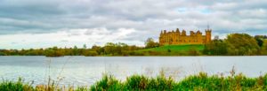 a long rectangular image split into four parts. a small sliver on the bottom is some tall bright green grass. Then a lake takes up the rest of the bottom half the photo before more land split the lake from the sky. On this bit of land sits an old school castle. The clouds in the sky take up the remainder of the photo