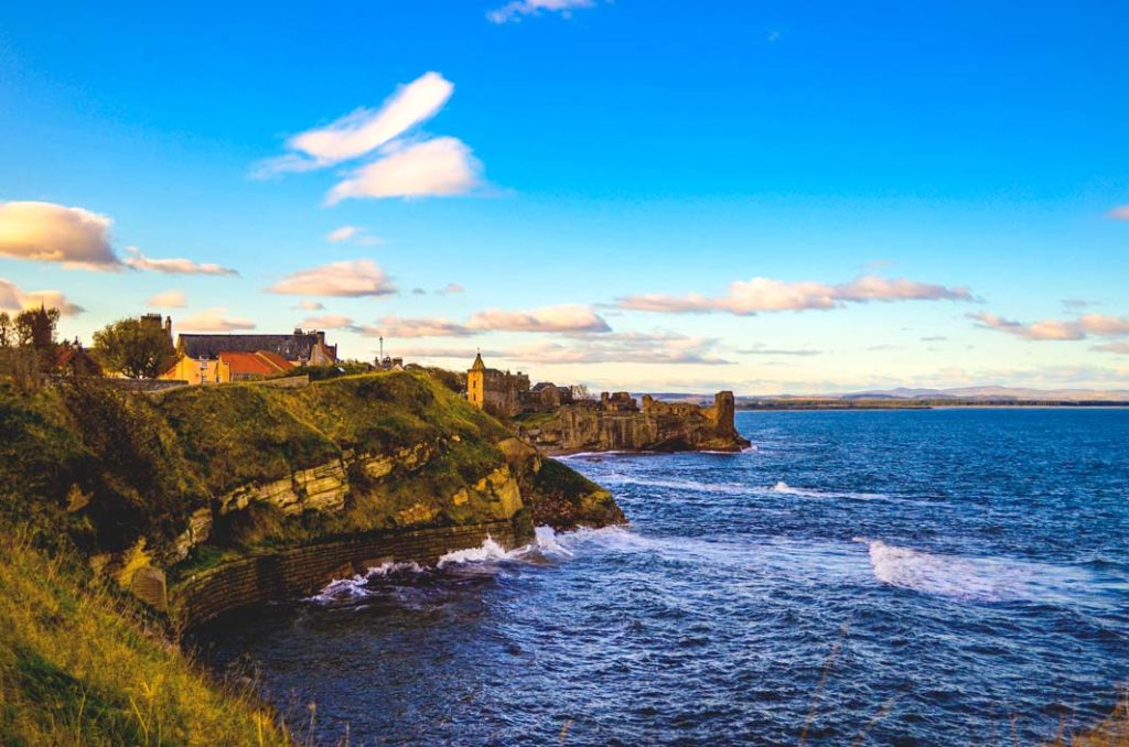 St Andrews cliffside views during golden hour. The rocks are cascading down in the rough see below as it hits the lowest part o the rock wall. The cliffs created two alcoves. On the farthest one you can slightly see the castler uins. 