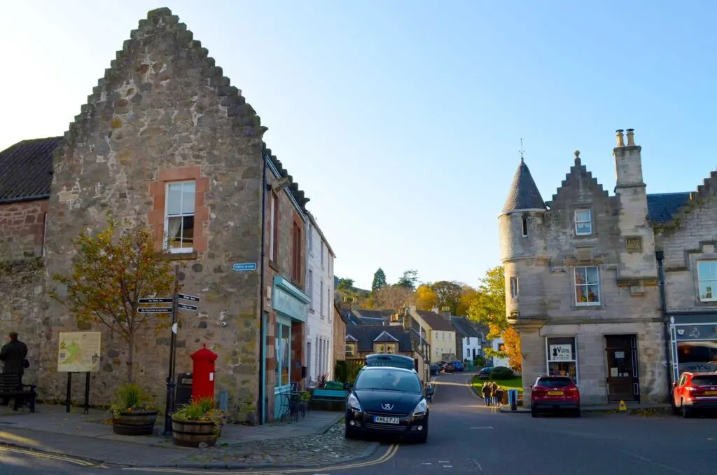 flakland village in scotland street corner. There is a car parked on the road in front of a two story stone house. It has a tall triangle roof. Another three story stone house faces you , it has a turret on the side a chimney connects it to the adjacent house. There are storefronts on the first level.