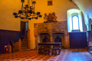 inside doune castle. It's complete with all four walls, a floor, and ceiling! The inside is fully done, red and white tiled floors . The walls are finished, and painted yellow. There's a double stone fireplace on the back wall, with the Scottish emblem above (a unicorn and a lion). Next to the fireplace is the only window in the room, it's completely white from the light shining in from the outside, giving the only light to the room. Hanging from the ceiling is an old iron or metal light figure, but it's not lit. The only way out of the room is a narrow wooden staircase leading you up.