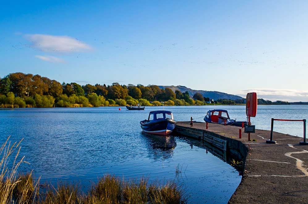 two boats are attached to a permanent paved dock in the lake. One boat faces you, while the other one on the opposite side of the dock, faces outward towards a completely tree covered island in the middle of the lake. 