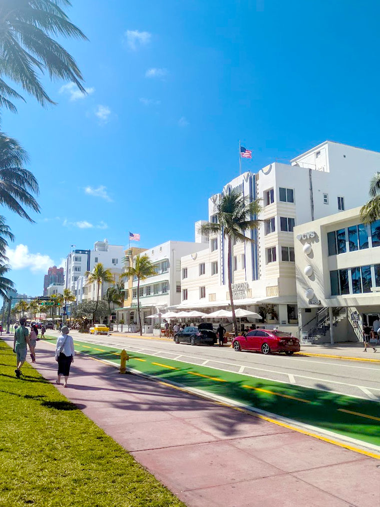 a bright sunny day in the art deco disctrict in south beach miami. People walk along the red sidewalk, sandwiched by grass on either side. You can along see the top corners of the palm trees that hang above. On the right side is the street, with some parked cars, and short art deco buildings, painted white with pastel accent colours. There are america flags blowing in the wind of the rooftops of two of them .