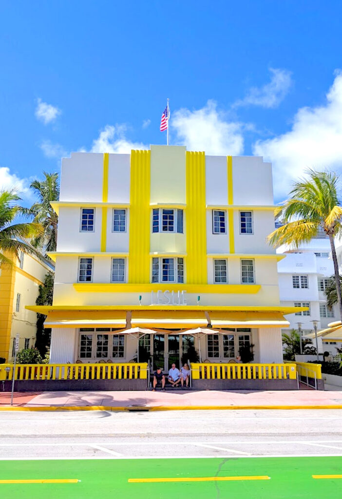 a vibrant happy photo with popping colours of an art deco hotel in south beach Miami in the art deco district. the hotel is onyl three stories tall, it's white and a lemon yellow paint with an awning shadowing the first floor. Three people sit on the steps leading to the hotel escaping the sun. There's a palm tree on either side of the hotel, both bending towards the left, from the wind. There are bright white clouds behind the hotel, but the rest of the sky is bright blue.